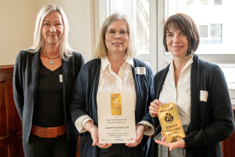 Drei Frauen stehen nebeneinander und schauen in die Kamera, die mittlere hält eine Urkunde in der Hand.